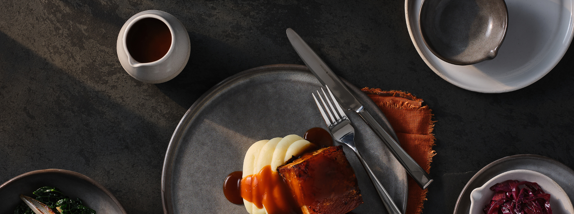 Pork belly on mashed potatoes on a deep grey glazed plate surrounded by grey and white crockery, some with a distinct indent, mimicking the thumb mark of the potter for an eye catching design.
